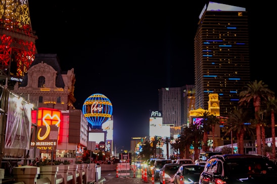 Bright and colorful Las Vegas cityscape at night with illuminated buildings and hotels. Neon signs and billboards light up the street. Cars are lined up on the road while palm trees are visible along the side.