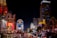 Bright and colorful Las Vegas cityscape at night with illuminated buildings and hotels. Neon signs and billboards light up the street. Cars are lined up on the road while palm trees are visible along the side.