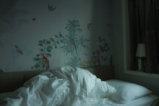 A rumpled white duvet and pillows are on a bed against a wall adorned with intricate nature-themed wallpaper featuring birds and plants. On the bedside, there's a lamp and dark curtains are drawn to one side.