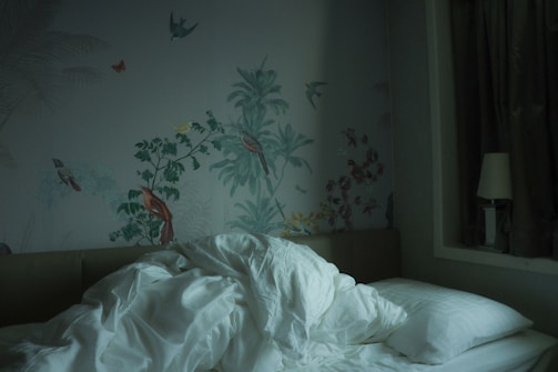 A rumpled white duvet and pillows are on a bed against a wall adorned with intricate nature-themed wallpaper featuring birds and plants. On the bedside, there's a lamp and dark curtains are drawn to one side.