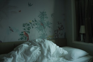 A rumpled white duvet and pillows are on a bed against a wall adorned with intricate nature-themed wallpaper featuring birds and plants. On the bedside, there's a lamp and dark curtains are drawn to one side.