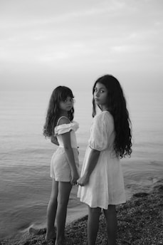 Two young girls with long hair stand by the shore, holding hands and looking towards the camera. They are dressed in light-colored summer clothing and the scene is captured in black and white, with calm sea waters in the background under a cloudy sky.