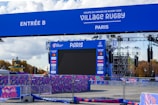 A large outdoor screen is set up for the Rugby World Cup 2023 event in Paris. The setup includes a blue archway marked 'Entrée B' and various banners with sponsor logos, surrounded by metal barriers. There are structures for lighting and sound, and trees are visible in the background under a partly cloudy sky.