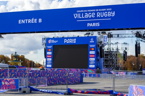 A large outdoor screen is set up for the Rugby World Cup 2023 event in Paris. The setup includes a blue archway marked 'Entrée B' and various banners with sponsor logos, surrounded by metal barriers. There are structures for lighting and sound, and trees are visible in the background under a partly cloudy sky.