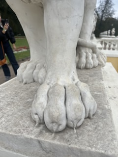 A close-up view of a marble statue's paw, showcasing the detailed craftsmanship of the sculpture. The texture of the marble is smooth with subtle vein patterns, and the statue stands on a marble base. A person in the background is taking a photo with a smartphone, and there are trees and ornamental gardens visible.