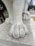 A close-up view of a marble statue's paw, showcasing the detailed craftsmanship of the sculpture. The texture of the marble is smooth with subtle vein patterns, and the statue stands on a marble base. A person in the background is taking a photo with a smartphone, and there are trees and ornamental gardens visible.