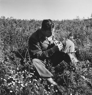 A person is sitting in a field of tall grass and wildflowers, intently examining an object in their hands. The person is wearing a military-style uniform and appears to be focused on their task.