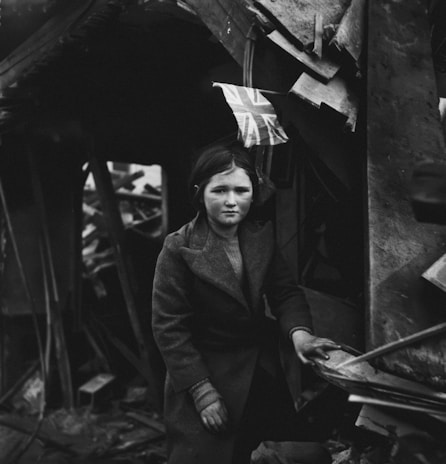 A young girl in a coat stands amid rubble and debris, holding onto a broken structure. She appears somber and is surrounded by damaged objects and a small Union Jack flag.