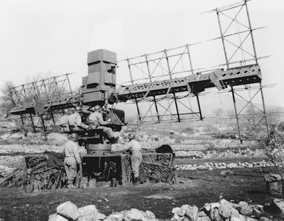 A group of soldiers operating a large radar machine in an outdoor, rural setting. The radar structure is sizable with multiple metal components, supported by a sturdy base and surrounded by camouflage netting. The soldiers are wearing military uniforms and helmets, and some are interacting with the equipment, suggesting a military exercise or operation during wartime.
