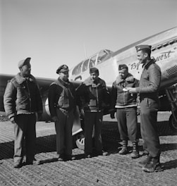 Five men wearing military uniforms stand in front of a fighter aircraft. They appear to be engaged in a conversation, with one person holding a notepad or document. The aircraft is a vintage model, suggesting a historical context.