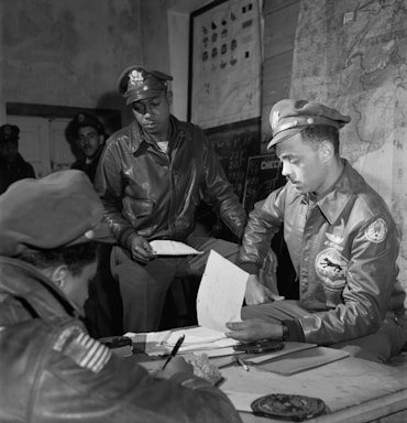 Several men in military uniforms are gathered around a table in a room that features maps and charts. They appear to be engaged in a meeting or briefing, with one individual holding a piece of paper while others write notes. The environment conveys a sense of focus and seriousness.