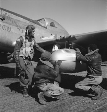 Three individuals, dressed in flight gear and leather jackets, work together on a military aircraft. One stands while two crouch to inspect a bomb or fuel tank under the wing. The airplane displays nose art and appears to be from the WWII era.