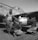 Three individuals, dressed in flight gear and leather jackets, work together on a military aircraft. One stands while two crouch to inspect a bomb or fuel tank under the wing. The airplane displays nose art and appears to be from the WWII era.