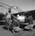 Three individuals, dressed in flight gear and leather jackets, work together on a military aircraft. One stands while two crouch to inspect a bomb or fuel tank under the wing. The airplane displays nose art and appears to be from the WWII era.