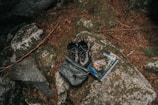 A pair of outdoor hiking shoes and a backpack are placed on a large rock surrounded by natural elements like pine needles and stones. Beside them is a guidebook or map with an outdoor adventure theme.