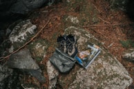 A pair of outdoor hiking shoes and a backpack are placed on a large rock surrounded by natural elements like pine needles and stones. Beside them is a guidebook or map with an outdoor adventure theme.
