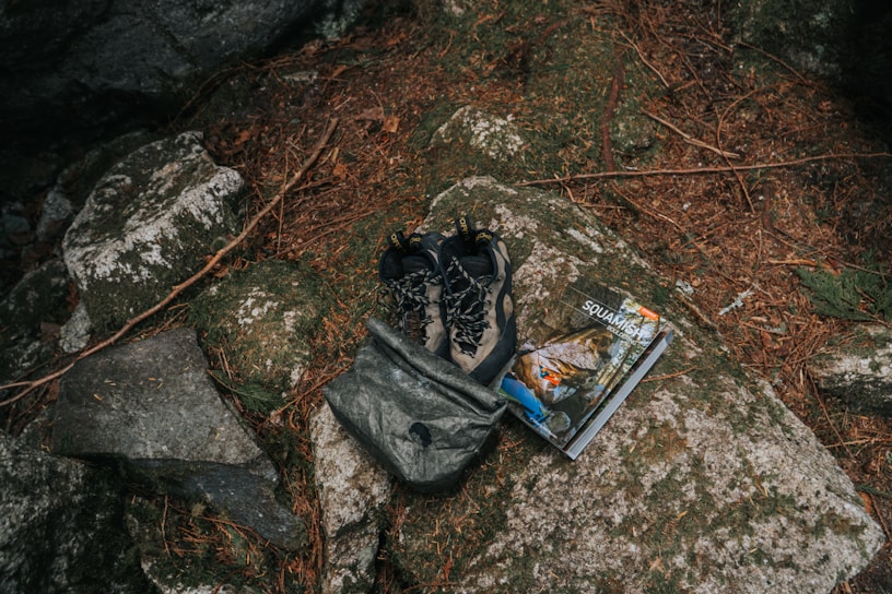 A pair of outdoor hiking shoes and a backpack are placed on a large rock surrounded by natural elements like pine needles and stones. Beside them is a guidebook or map with an outdoor adventure theme.