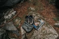 A pair of outdoor hiking shoes and a backpack are placed on a large rock surrounded by natural elements like pine needles and stones. Beside them is a guidebook or map with an outdoor adventure theme.
