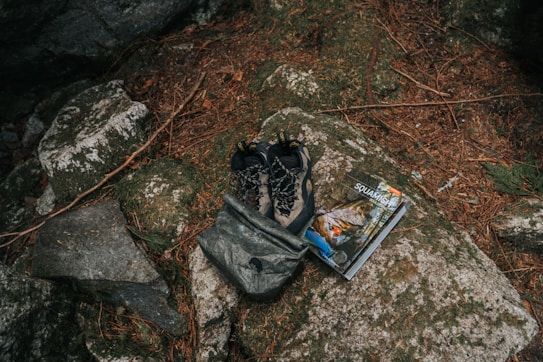A pair of outdoor hiking shoes and a backpack are placed on a large rock surrounded by natural elements like pine needles and stones. Beside them is a guidebook or map with an outdoor adventure theme.
