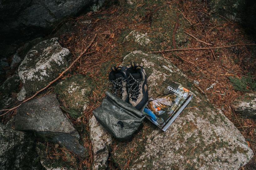 A pair of outdoor hiking shoes and a backpack are placed on a large rock surrounded by natural elements like pine needles and stones. Beside them is a guidebook or map with an outdoor adventure theme.