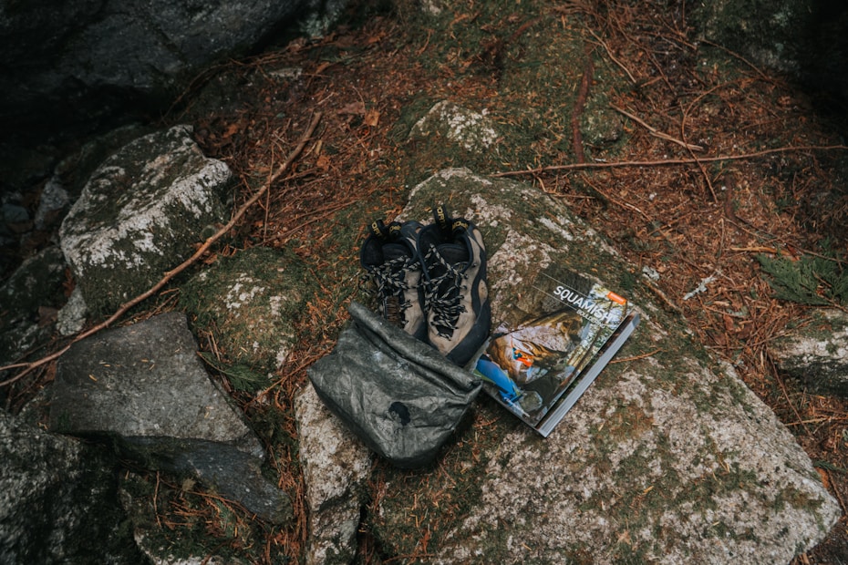A pair of outdoor hiking shoes and a backpack are placed on a large rock surrounded by natural elements like pine needles and stones. Beside them is a guidebook or map with an outdoor adventure theme.