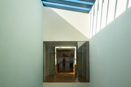 A modern architectural setting featuring a glass ceiling that allows natural light to pour into a corridor. The focal point is a large window leading to another room with a person visible in the reflection, capturing a background view of a traditional rooftop. The play of light and shadows creates geometric patterns on the walls.