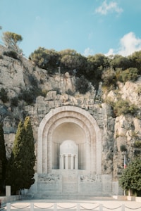 A monumental structure is built into a rocky hillside, featuring a large arch and a central columned rotunda. Tall cypress trees flank the structure on one side, while a French flag is displayed to its right. The surrounding area includes lush greenery and rugged rock formations under a clear blue sky.