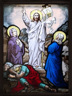 A stained glass artwork depicting a central figure in a white robe, holding a staff with a flag, surrounded by two kneeling figures with halos and two sleeping figures. The scene appears to be on a rocky landscape.