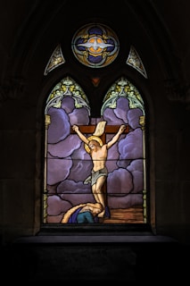 A stained glass window featuring a depiction of the crucifixion, with a religious figure on a cross set against a backdrop of purple and blue clouds. The window is framed with ornate designs and symbols.