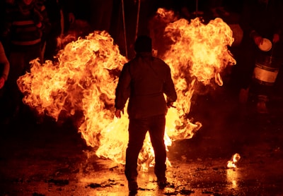 A person stands in front of a large, intense fire that creates dramatic lighting, with flames prominently rising. Other people are partially visible in the background, observing the scene.