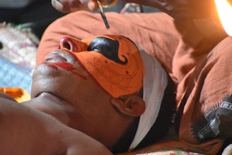 A person is lying down while someone applies intricate face paint, featuring bright orange and black patterns, highlighting cultural or ceremonial artwork. The focus is on the detailed brushwork and vivid colors against the skin, showcasing a traditional practice.