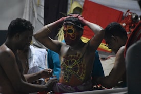 A person with intricate body paint and a vibrant face mask stands with arms raised, surrounded by two individuals assisting in the adornment process. The setting features vivid colors and decorative elements suggestive of a traditional ritual or ceremony.