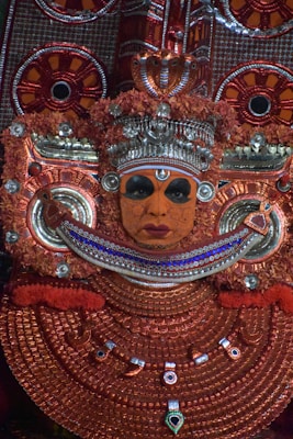 Intricately detailed traditional costume and makeup featuring vivid orange and red colors, adorned with metallic embellishments. The face is painted with elaborate designs, and the headpiece includes ornate floral elements and jewels.