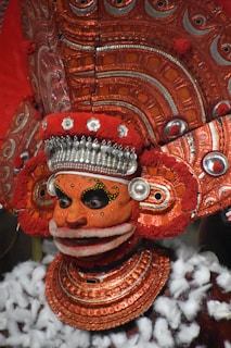 A highly ornate and colorful traditional costume featuring a mask with intricate designs and patterns. The headpiece and collar are elaborately decorated with metallic details and rich, textured materials. The colors are predominantly red and orange, with accents of white and silver. The overall appearance is ceremonial and artistic, indicative of cultural or ritualistic significance.