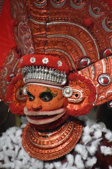 A highly ornate and colorful traditional costume featuring a mask with intricate designs and patterns. The headpiece and collar are elaborately decorated with metallic details and rich, textured materials. The colors are predominantly red and orange, with accents of white and silver. The overall appearance is ceremonial and artistic, indicative of cultural or ritualistic significance.