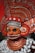 A highly ornate and colorful traditional costume featuring a mask with intricate designs and patterns. The headpiece and collar are elaborately decorated with metallic details and rich, textured materials. The colors are predominantly red and orange, with accents of white and silver. The overall appearance is ceremonial and artistic, indicative of cultural or ritualistic significance.