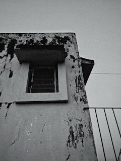 An aged, weathered building facade with peeling paint and visible black mold or grime marks. A single window with metal bars is present, framed by the worn structure. There is a portion of a railing to the right, suggesting the presence of a balcony or stairway.