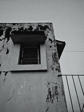 An aged, weathered building facade with peeling paint and visible black mold or grime marks. A single window with metal bars is present, framed by the worn structure. There is a portion of a railing to the right, suggesting the presence of a balcony or stairway.