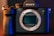 A professional digital camera with a sleek black body design and prominent grip. The brand logo is visible on top, and the camera's lens is removed, exposing the sensor. The scene is lit with a combination of warm and cool tones, highlighting the detailed textures of the camera.