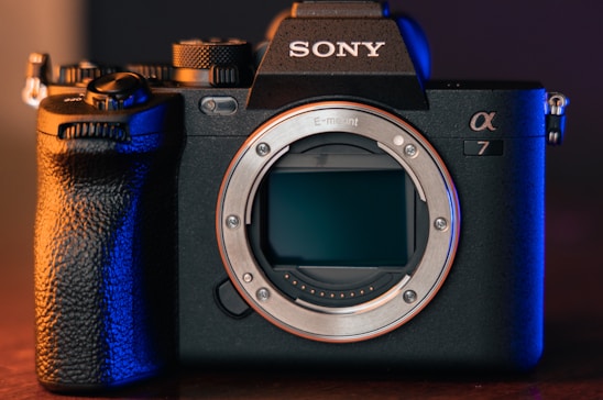 A professional digital camera with a sleek black body design and prominent grip. The brand logo is visible on top, and the camera's lens is removed, exposing the sensor. The scene is lit with a combination of warm and cool tones, highlighting the detailed textures of the camera.