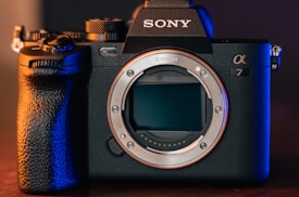 A professional digital camera with a sleek black body design and prominent grip. The brand logo is visible on top, and the camera's lens is removed, exposing the sensor. The scene is lit with a combination of warm and cool tones, highlighting the detailed textures of the camera.