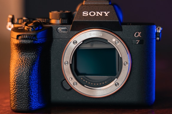 A professional digital camera with a sleek black body design and prominent grip. The brand logo is visible on top, and the camera's lens is removed, exposing the sensor. The scene is lit with a combination of warm and cool tones, highlighting the detailed textures of the camera.