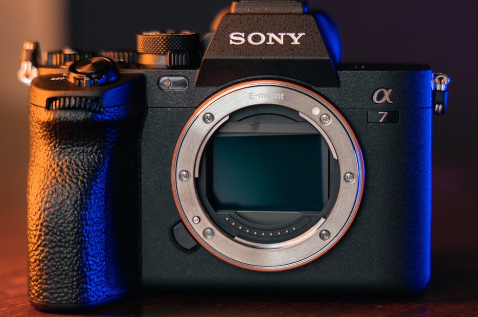 A professional digital camera with a sleek black body design and prominent grip. The brand logo is visible on top, and the camera's lens is removed, exposing the sensor. The scene is lit with a combination of warm and cool tones, highlighting the detailed textures of the camera.
