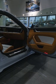 The interior of a luxury car showroom features an open car door with brown leather and fabric upholstery. Bright showroom lights illuminate the sleek design, while large windows allow natural daylight to stream in. A framed photograph of a blue sports car is displayed on the wall above.