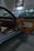 The interior of a luxury car showroom features an open car door with brown leather and fabric upholstery. Bright showroom lights illuminate the sleek design, while large windows allow natural daylight to stream in. A framed photograph of a blue sports car is displayed on the wall above.