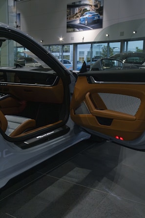 The interior of a luxury car showroom features an open car door with brown leather and fabric upholstery. Bright showroom lights illuminate the sleek design, while large windows allow natural daylight to stream in. A framed photograph of a blue sports car is displayed on the wall above.