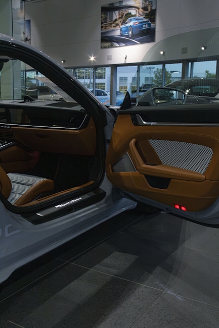 The interior of a luxury car showroom features an open car door with brown leather and fabric upholstery. Bright showroom lights illuminate the sleek design, while large windows allow natural daylight to stream in. A framed photograph of a blue sports car is displayed on the wall above.