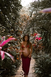 A pregnant woman walks along a brick path surrounded by lush tropical foliage. The greenery includes palm leaves and plants with vibrant red and pink flowers. She is wearing a maroon skirt and a red bikini top, and the surroundings are dense with vegetation, creating a serene, natural atmosphere.