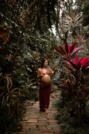 A pregnant woman walks along a cobblestone path surrounded by lush, tropical foliage. She wears a red wrap skirt and a bikini top, gently touching the leaves of tall plants on either side.