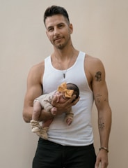 A man with tattoos stands against a plain background, wearing a white tank top and black jeans. He is holding a small baby adorned with a large yellow bow on its head. The baby is dressed in a beige outfit and small booties, and the man has a gentle expression.